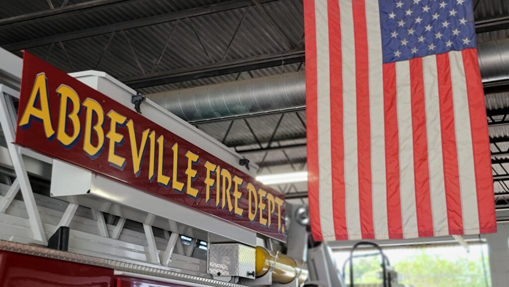 Interior of the Abbeville Fire Department showing a ladder truck with “Abbeville Fire Dept.” signage and a large American flag hanging from the station ceiling.