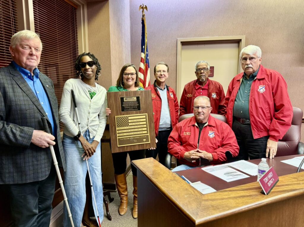 Mayor and members of the Louisiana/Mississippi Blinded Veterans Association stand with city officials in the City Council chamber holding the Braille American Flag plaque during a presentation.